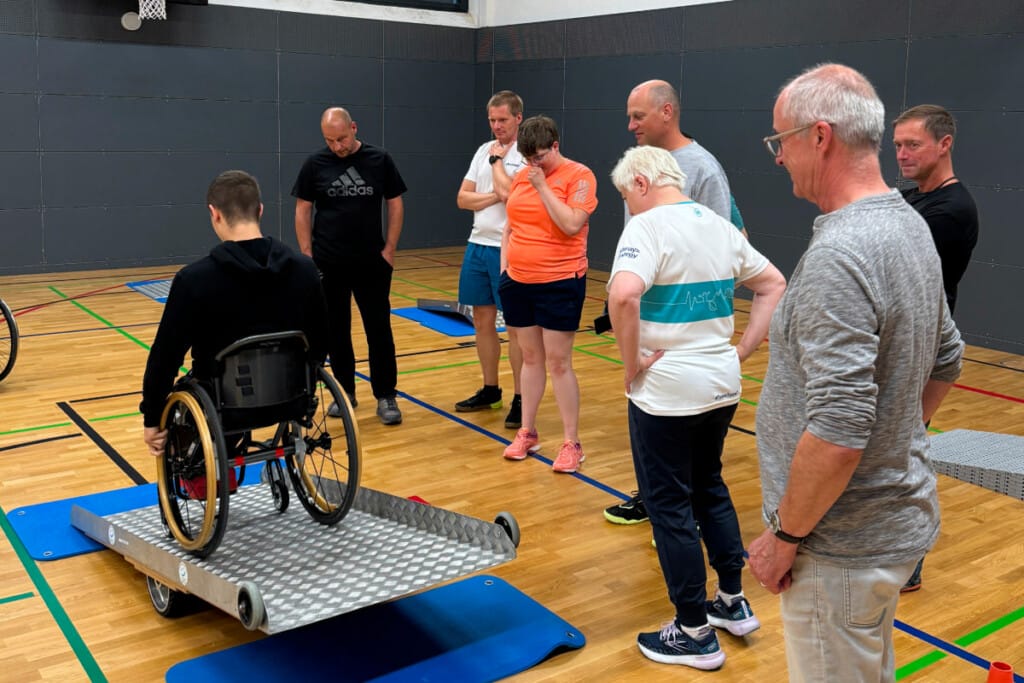 Person fährt mit Rollstuhl über Rampe in Sporthalle und weitere schauen dabei zu