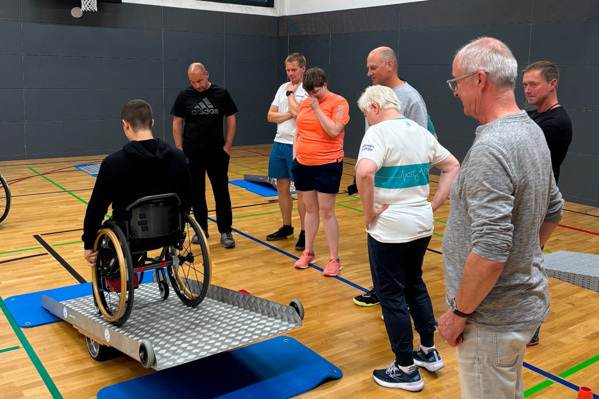 Person fährt mit Rollstuhl über Rampe in Sporthalle und weitere schauen dabei zu