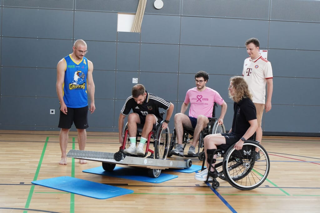 Personen fahren mit Rollstuhl über eine Rampe die in Sporthalle aufgebaut ist, weitere schauen zu