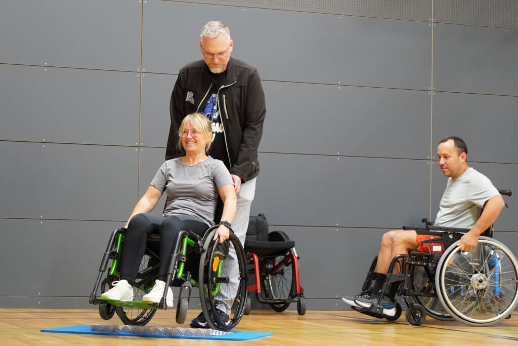 Person im Rollstuhl wird von weiterer Person über Hindernis geschoben