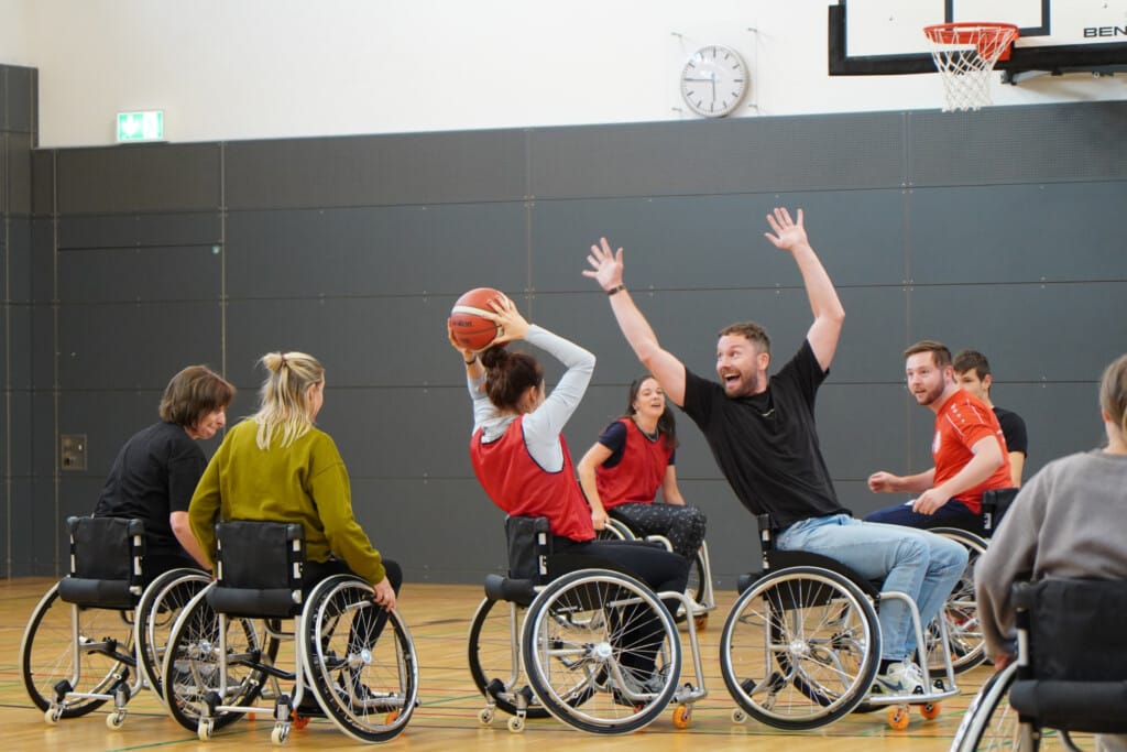 Personen spielen Rollstuhlbasketball, dabei wirft eine auf den Korb und wird jemand anderem geblockt
