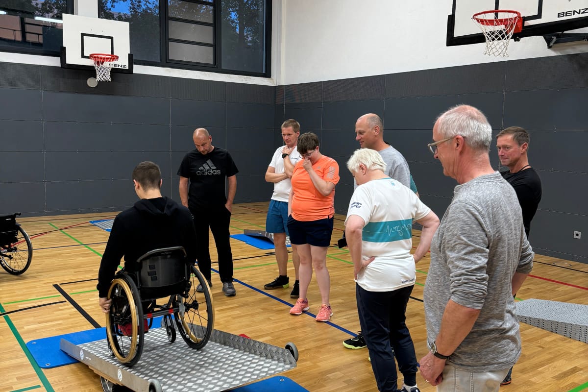 Person mit Rollstuhl fährt in Turnhalle über Rampe, weitere Personen stehen daneben und schauen zu