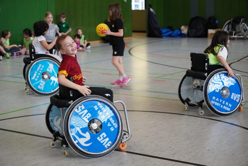 Kinder fahren im Rollstuhl durch Sporthalle