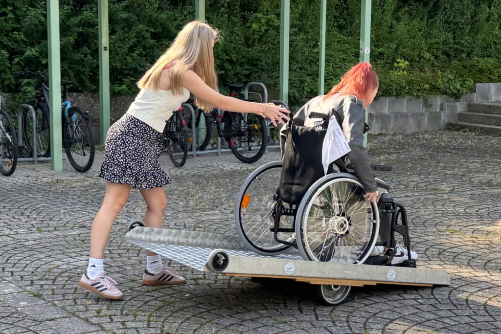 Schülerin im Rollstuhl fährt auf Pflasterstein über Rampe, eine weitere Schülerin hilft ihr dabei