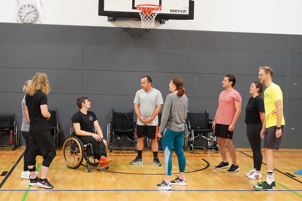 Teilnehmende eines Firmenevents beim RSV Bayreuth erhalten in der Sporthalle eine Einführung in den Rollstuhlbasketball und bereiten sich gemeinsam auf die nächsten Übungen vor.